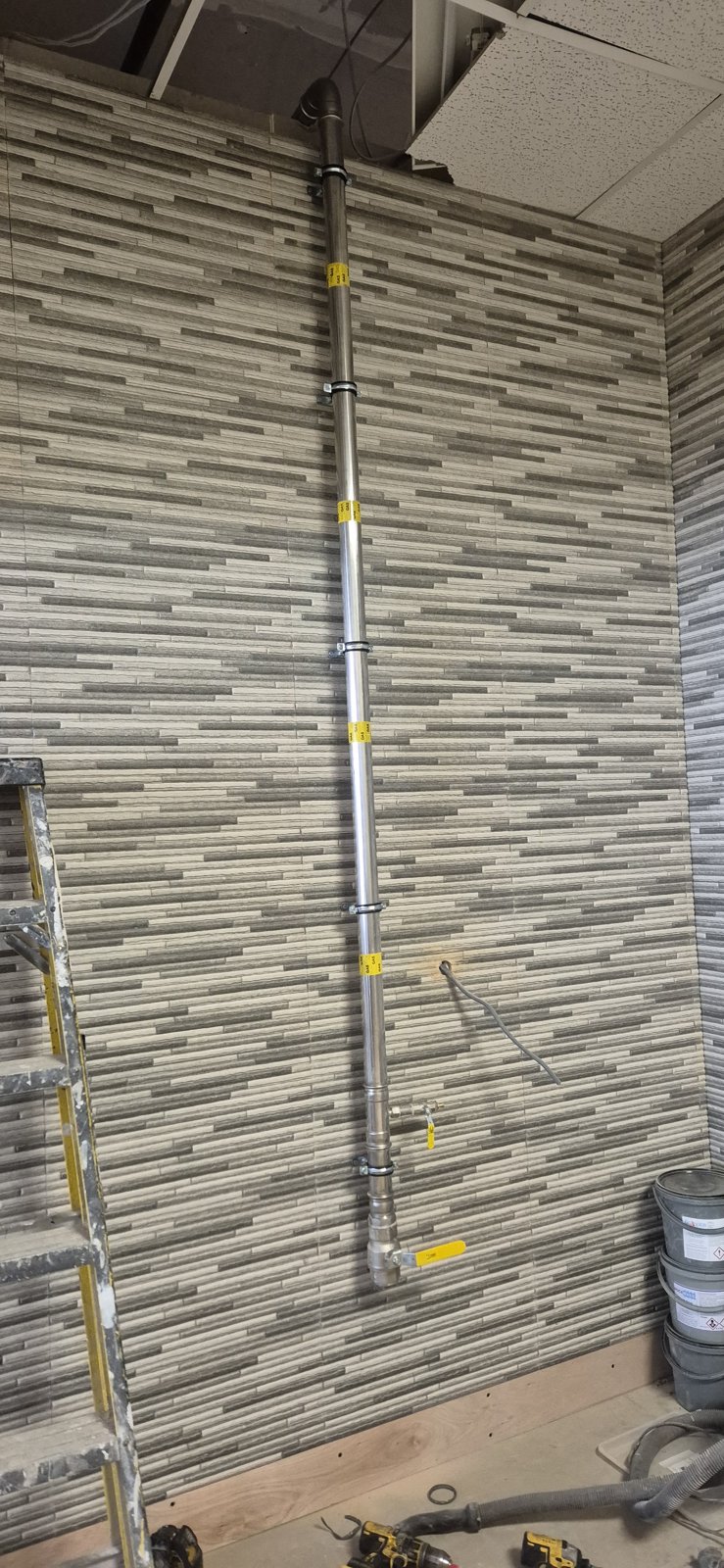 A stainless steel gas supply pipe with yellow gas labels and an isolation valve on a tiled commercial wall